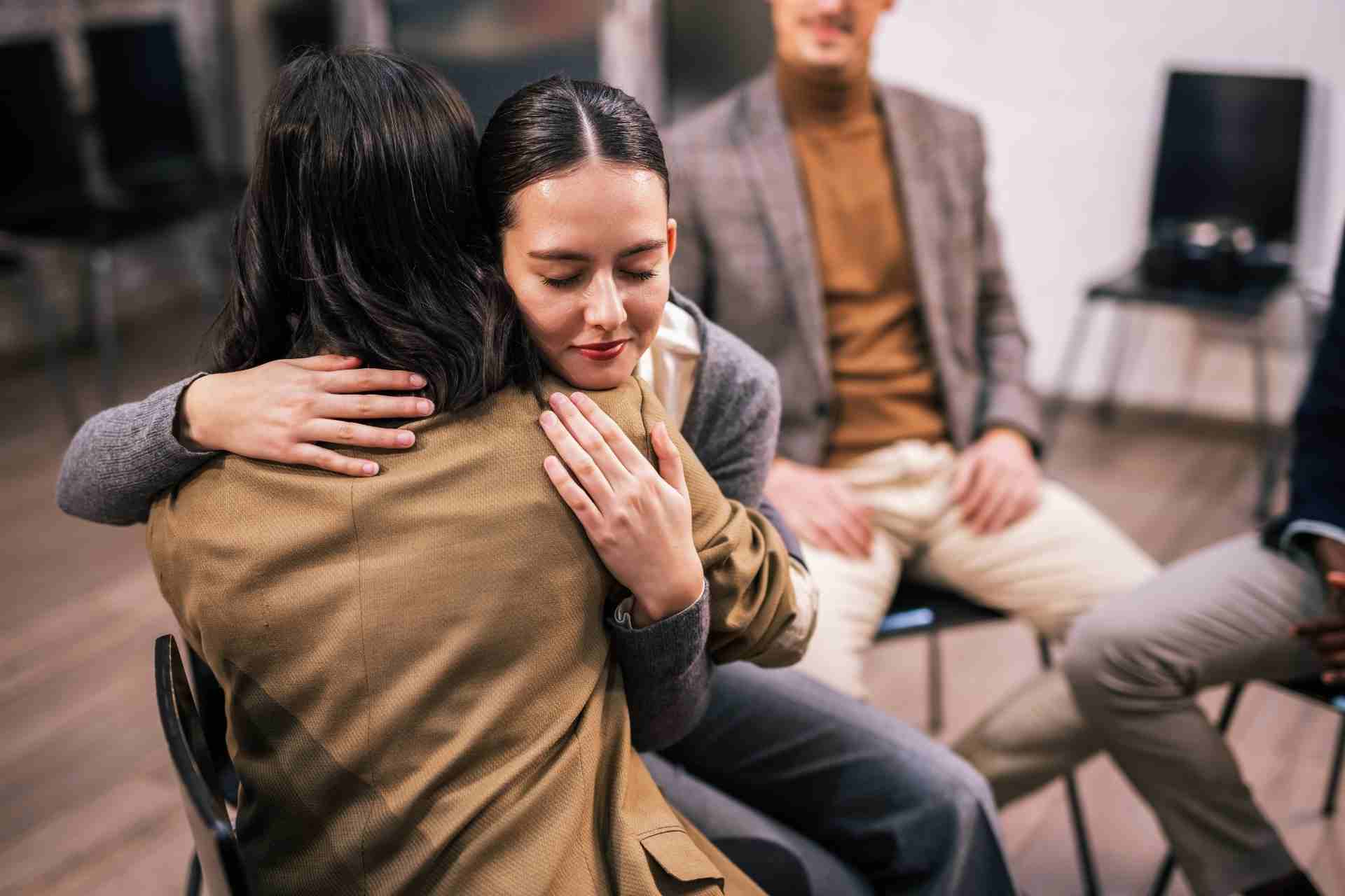 Two participants embrace during a mental health intensive outpatient program (IOP) group therapy session while other group members provide support in the background.