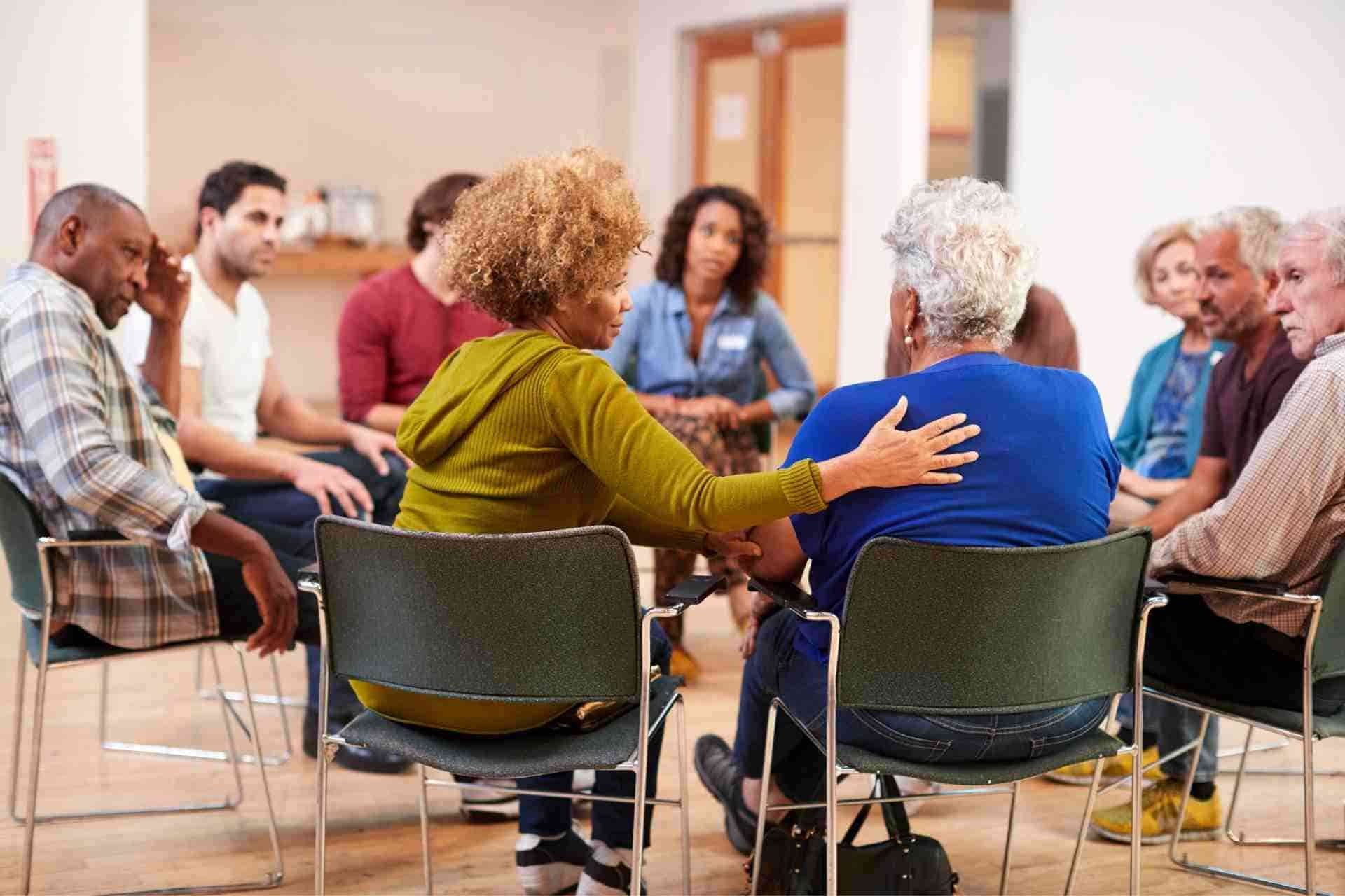 A participant comforts another during a therapist-led mental health group therapy session while others listen attentively.