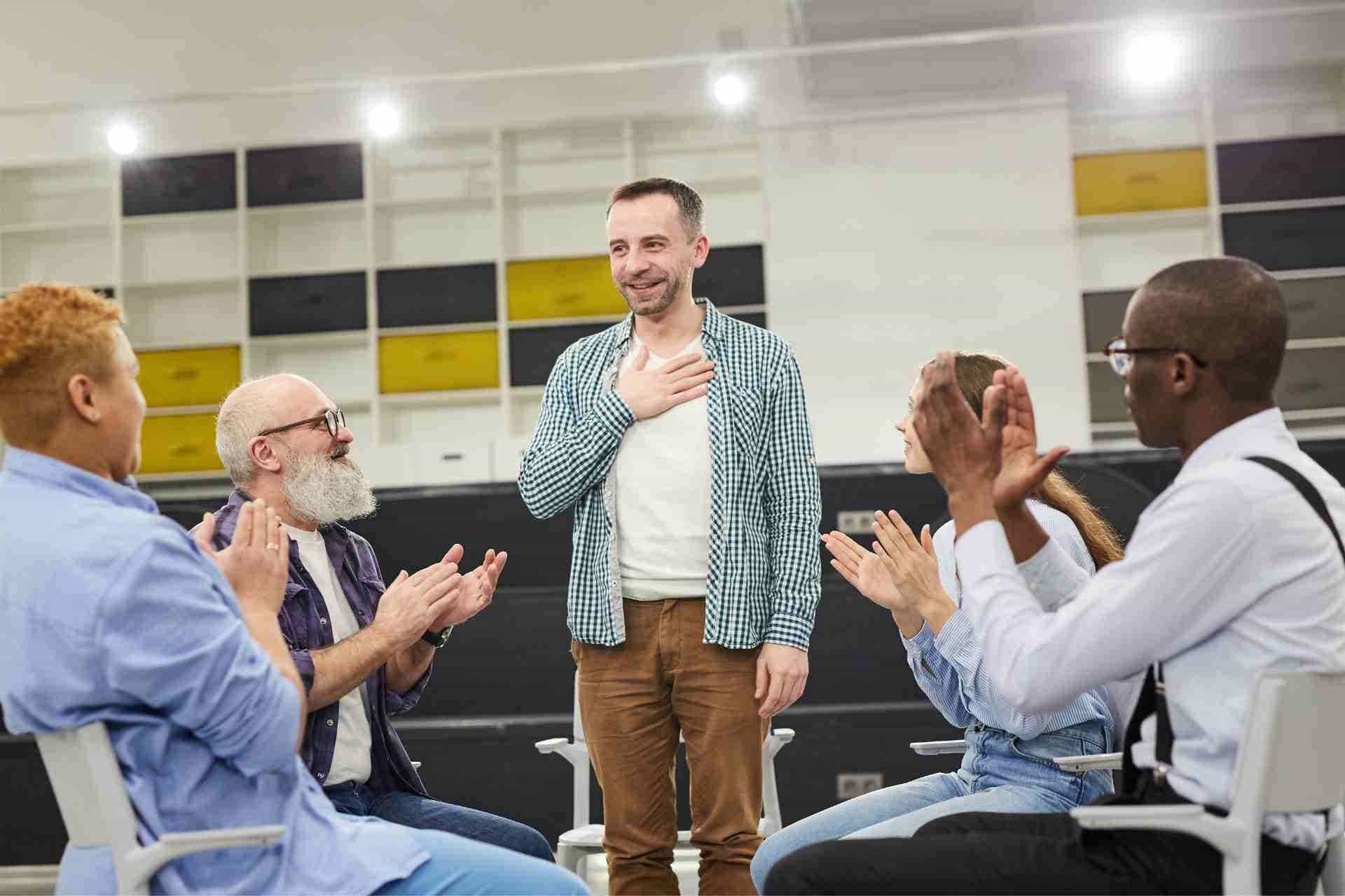 Mental health therapy group applauds as a participant shares recovery progress during a counseling session.