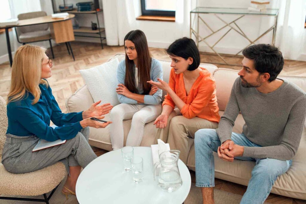 Female therapist facilitating a family therapy session with a teenage girl and two adults seated together on a couch.
